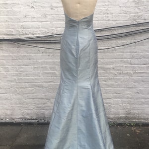 May include: A light blue, floor-length gown with a halter neckline and a mermaid silhouette. The dress is displayed on a mannequin, showcasing its open back and elegant design. The fabric has a subtle sheen.