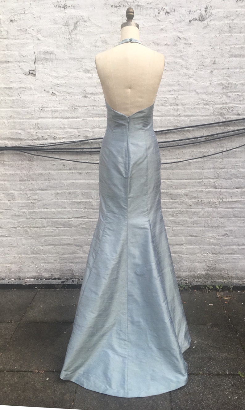 May include: A light blue, floor-length gown with a halter neckline and a mermaid silhouette. The dress is displayed on a mannequin, showcasing its open back and elegant design. The fabric has a subtle sheen.