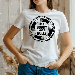 Puede incluir: Camiseta blanca con un gráfico circular que dice "Be a Buddy Not a Bully". El diseño presenta manos que rodean el texto, con palabras como "Unity", "Peace" e "Inclusion" en el borde. La camiseta es de manga corta.