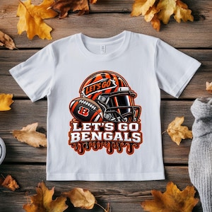 Puede incluir: Una camiseta blanca de manga corta con un gráfico de un casco de fútbol americano y un balón con rayas de tigre naranjas y negras. El diseño incluye el texto "LET'S GO" y "LET'S GO BENGALS" en letras blancas en negrita con borde negro.