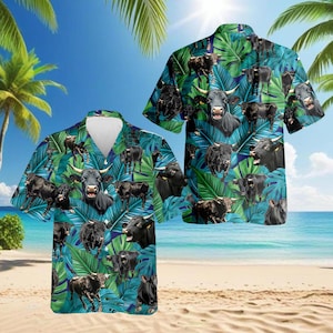 Op de afbeelding: Een shirt met korte mouwen met een tropisch ontwerp. Het shirt heeft zwarte stieren en groene bladeren op een blauwe achtergrond. Het shirt wordt getoond op een strand met palmbomen en de oceaan op de achtergrond. Het shirt heeft een klassieke kraag en een relaxte pasvorm.