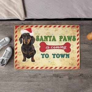 May include: A rectangular doormat featuring a dachshund wearing a Santa hat. The mat displays the text "SANTA PAWS is coming TO TOWN" in green and red. The background is beige with paw prints and snowflakes.