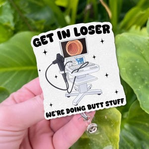 以下が含まれることがあります： 「GET IN LOSER」の文字と医療機器のイラスト、そして「WE'RE DOING BUTT STUFF」の文字が描かれた白いバッジリール。 バッジリールには透明なプラスチック製の格納式コードと金属製のクリップが付いています。