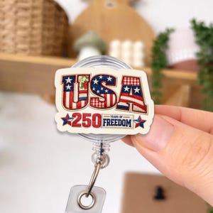 May include: A badge reel with a patriotic design. The badge reel features the word "USA" in red, white, and blue, with stars and stripes. The text "250 YEARS OF FREEDOM" is also included.