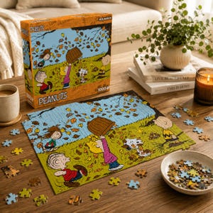 May include: A 1000-piece Peanuts jigsaw puzzle box and partially assembled puzzle featuring characters playing in autumn leaves. The scene shows Snoopy, Charlie Brown, and friends in a grassy field with falling leaves, matching the box art.