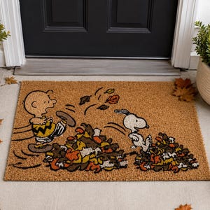 May include: A rectangular tan coir doormat featuring a graphic illustration of Charlie Brown and Snoopy playing in piles of colorful autumn leaves. The design shows the characters in motion, with scattered leaves floating in the air above the piles.