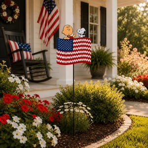 May include: A decorative American flag yard stake featuring Charlie Brown and Snoopy. The flag has a red and white zig-zag design with a blue star field. The yard stake is placed in a garden with red and white flowers.