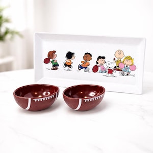 May include: A white rectangular serving platter and two maroon football-shaped bowls. The platter features a cartoon illustration of children playing football. The bowls have white stripes and stitching details.