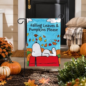 May include: A rectangular garden flag featuring a white cartoon dog lying on a red roof against a light blue sky. The text Falling Leaves & Pumpkins Please is printed in black above the dog, surrounded by colorful autumn leaves and orange pumpkins.