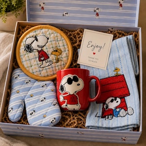 May include: A gift box containing a round quilted potholder with Snoopy and Woodstock, a blue and white striped oven mitt, a red mug featuring Joe Cool Snoopy, and a blue striped towel with Snoopy on a red doghouse, plus a card reading Enjoy! Thank you so much.