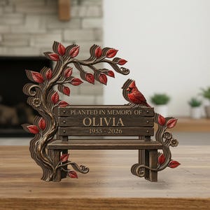 Könnte beinhalten: Ein dekoratives Gedenkbank-Ornament in brauner Holzoptik mit modellierten Ästen und roten Blättern. Ein roter Kardinalvogel sitzt auf der Rückenlehne, die mit dem Text PLANTED IN MEMORY OF OLIVIA 1955-2026 graviert ist.
