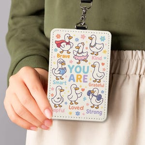 May include: A white lanyard ID badge with cartoon ducks and positive affirmations. The badge has a black lanyard and a silver clasp. The ducks are shown with accessories and the words "YOU ARE" are in the centre.