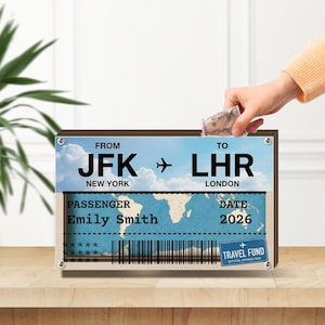 Può includere: Un salvadanaio rettangolare per fondi di viaggio, progettato come un biglietto aereo. Il biglietto mostra "JFK" a "LHR", "New York" a "Londra", "Emily Smith" e "2026". Una mano inserisce denaro nella fessura.