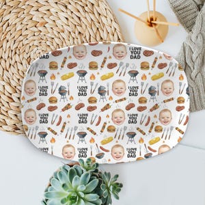 Peut inclure: Plat ovale blanc avec un motif répété de visages de bébés, de barbecues et d'aliments. Les mots "I LOVE YOU DAD" sont également imprimés. Le plat est entouré d'un dessous de plat tissé, d'une plante succulente et d'une couverture en tricot grise.