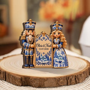 May include: A wooden ornament featuring a nutcracker and a female figure in blue and white winter-themed attire, holding a central plaque with the text Olivia & Noah and 12.07.2026, decorated with snowflake patterns and a snowy landscape scene.
