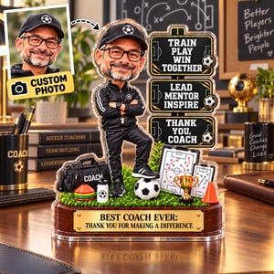 May include: A custom bobblehead figure of a coach wearing a black tracksuit, cap, and glasses, standing on a green base with a football, bag, and cones. Beside the figure are signs reading TRAIN PLAY WIN TOGETHER, LEAD MENTOR INSPIRE, THANK YOU, COACH, and BEST COACH EVER: THANK YOU FOR MAKING A DIFFERENCE.