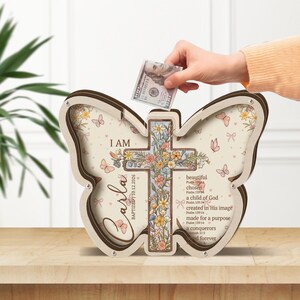 Puede incluir: Una hucha de madera con forma de mariposa y diseño de cruz. El banco presenta detalles florales, el nombre "Carla" y el texto "I AM". Una mano introduce un billete de 100 euros en la ranura. El banco está sobre una superficie de madera.