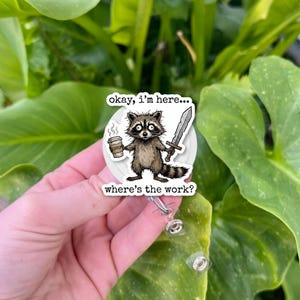 May include: A circular badge reel featuring a cartoon raccoon illustration holding a coffee cup and a sword. The text reads, "okay, i'm here... where's the work?" The design is printed on a white background with a clear plastic strap and metal snap.