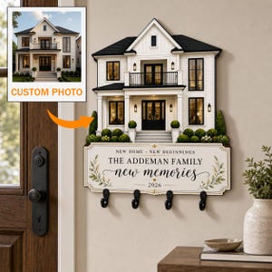 May include: A wall-mounted key holder featuring a three-dimensional model of a two-story white house with a black roof. Below the house, a rectangular sign displays the text NEW HOME - NEW BEGINNINGS, THE ADDEMAN FAMILY, new memories, 2026.