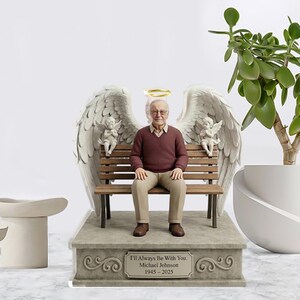 May include: A commemorative figurine featuring a man in a maroon jumper and tan trousers sitting on a wooden bench. Large white angel wings and two small cherubs flank the figure, with a gold halo above. The base bears the text I'll Always Be With You. Michael Johnson 1945-2025.