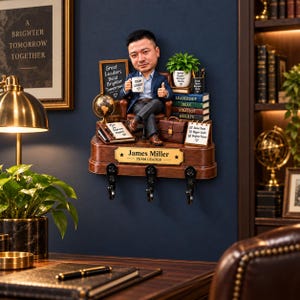 May include: A custom bobblehead figurine of a person in a suit sitting in a chair, mounted on a wooden wall shelf with three black hooks. The shelf features a nameplate reading James Miller Team Leader and various miniature office accessories.