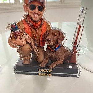 May include: A custom acrylic cutout display featuring a smiling person wearing a cap, sunglasses, and an orange vest, kneeling while holding a pheasant. Beside them sits a brown dog with a colourful collar, and a rifle stands upright on the base labelled DREW.