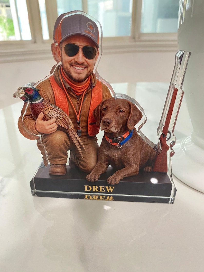May include: A custom acrylic cutout display featuring a smiling person wearing a cap, sunglasses, and an orange vest, kneeling while holding a pheasant. Beside them sits a brown dog with a colourful collar, and a rifle stands upright on the base labelled DREW.