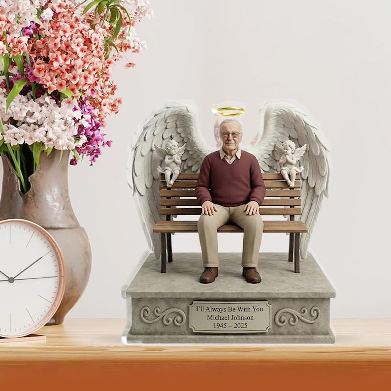 May include: A commemorative figurine featuring an elderly man with glasses wearing a maroon sweater and tan trousers, sitting on a wooden bench. The figure has large white angel wings, a gold halo, and two small cherubs. A plaque reads: I'll Always Be With You. Michael Johnson 1945-2025.