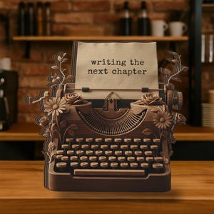 May include: A layered wooden typewriter sculpture featuring intricate floral carvings of roses and daisies on the sides. A paper insert at the top displays the black printed text "writing the next chapter" in a classic typewriter font style.