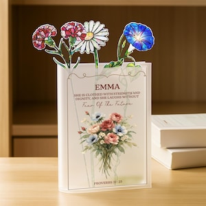 May include: A clear acrylic book stand with a floral design and the name "EMMA" printed on it. The stand features a bouquet of flowers and the text "SHE IS CLOTHED WITH STRENGTH AND DIGNITY, AND SHE LAUGHS WITHOUT Fear Of The Future".