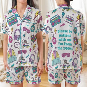 May include: White pajama set with a retro 1990s theme. The short-sleeved top and shorts feature colorful illustrations of boomboxes, roller skates, sunglasses, and the text "please be patient with me I'm from the 1900's". The set is trimmed with teal piping.