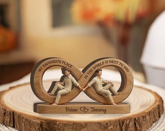 May include: A wooden infinity-shaped ornament featuring two figures sitting on the loops, holding hands. The text reads "MY FAVOURITE PLACE IN THE WORLD IS NEXT TO YOU" along the top, with "Brian" and "Tommy" separated by a heart on the base.