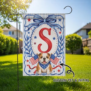 May include: A rectangular garden flag featuring a large red letter S centered within a decorative border. Below the letter, a brown and white bulldog wearing a blue star-patterned bandana is flanked by two small American flags and red ribbons.