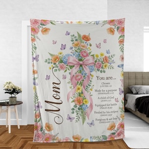 May include: A white blanket featuring a cross design made of colorful flowers and a pink bow. The word "Mom" is written on the left side. Text on the right side includes Bible verses about being chosen, made for a purpose, and forgiven.