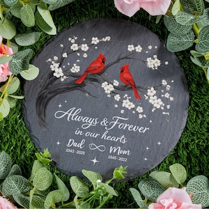Peut inclure: Une pierre tombale ronde et grise avec l'inscription "Always & Forever in our hearts", les noms "Dad" et "Mom" et des dates. Deux cardinaux rouges sont perchés sur une branche fleurie. Fleurs blanches et étoiles ornent le design.