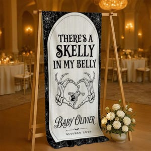 May include: A vertical banner with a cream-colored center and black borders featuring spiderweb patterns. The text reads THERE'S A SKELLY IN MY BELLY, BABY OLIVIER, and OCTOBER 2026. A graphic of two skeleton hands forming a heart shape surrounds a small skeleton.