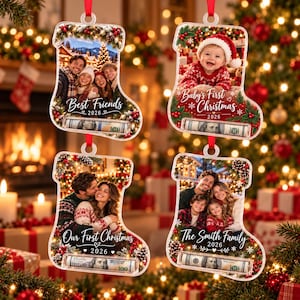 May include: Four clear acrylic stocking-shaped ornaments with red ribbons. Each features a photo, festive greenery, and the year 2026. A slot at the bottom holds a rolled 100 dollar bill. Text includes Best Friends, Baby's First Christmas, and Our First Christmas.