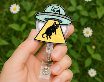 May include: A badge reel featuring a holographic UFO design with a black cat visible in the cockpit and a second black cat being abducted in a yellow holographic beam of light. The circular reel has a clear plastic strap with a metal snap button.