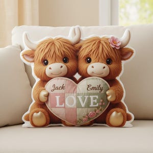 May include: A plush pillow shaped like two Highland cows holding a heart-shaped quilt. The quilt is pink and green with the names "Jack" and "Emily" and the word "LOVE". One cow has a pink flower.
