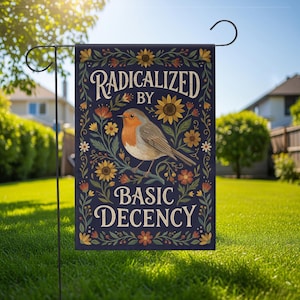 Op de afbeelding: Een marineblauwe tuinvaandel met een vogelillustratie en bloemen. De tekst luidt "RADICALIZED BY BASIC DECENCY". De vlag wordt buiten in een tuin getoond. De vlag is bevestigd aan een zwarte metalen paal.
