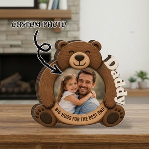 Könnte beinhalten: Ein hölzerner, bärenförmiger Fotorahmen mit einem Bild von Vater und Kind. Der Rahmen hat das Wort "Daddy" an der Seite und den Satz "Big Hugs for the Best Dad" unten. Der Text "Custom Photo" befindet sich über dem Rahmen.