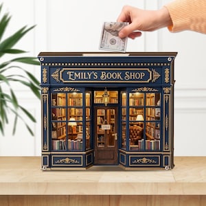 Può includere: Un modello in miniatura di una libreria chiamata Emily's Book Shop, caratterizzato da un esterno blu scuro con finiture dorate, ampie vetrine e un interno illuminato da una luce calda, con scaffali pieni di libri e una poltrona marrone.
