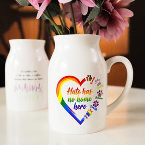 以下が含まれることがあります： 白いセラミック花瓶、虹色のハートのデザインと「Hate has no home here」の文字が特徴です。「Kindness」や「Diversity」などの足跡と単語も含まれています。花瓶にはピンクの花が入っています。