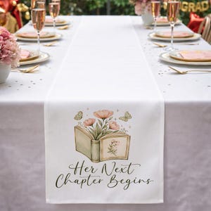 May include: White table runner with a floral and butterfly design. The runner features an open book illustration with pink flowers and the text "Her Next Chapter Begins". The runner is set on a white tablecloth with place settings.
