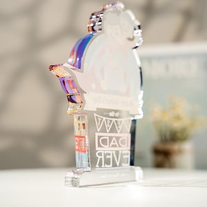 May include: A clear acrylic plaque with a die-cut top edge featuring a printed illustration of a person carrying another on their back. The text reads Happy Father's Day to the BEST DAD EVER. The edges of the acrylic display a reflective iridescent sheen.