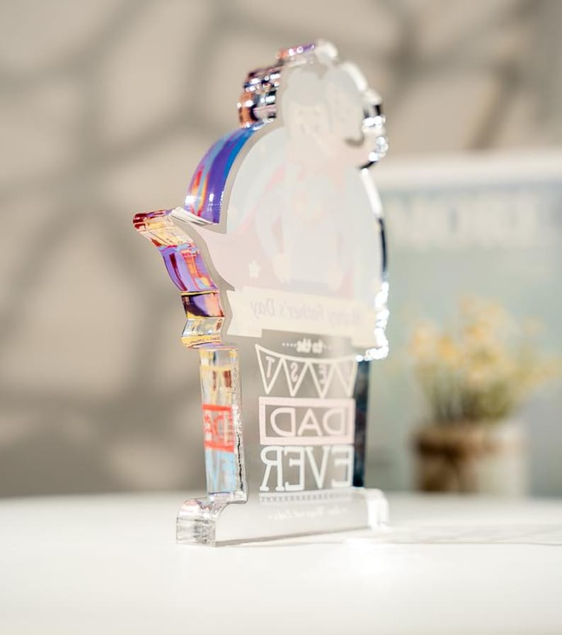 May include: A clear acrylic plaque with a die-cut top edge featuring a printed illustration of a person carrying another on their back. The text reads Happy Father's Day to the BEST DAD EVER. The edges of the acrylic display a reflective iridescent sheen.