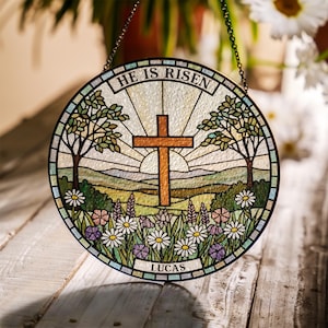 May include: Circular stained glass panel with a religious theme. It depicts a brown cross, a sunrise, trees, and flowers. The text "HE IS RISEN" is at the top, and "LUCAS" at the bottom. The panel is suspended by a chain.