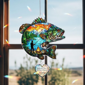 May include: A stained glass-style hanging ornament shaped like a fish, featuring a landscape with a person fishing by a campfire, mountains, and a sunset. A heart-shaped pendant hangs below with the text: Someone I love is Fishing in heaven, Henry, 1960-2026.