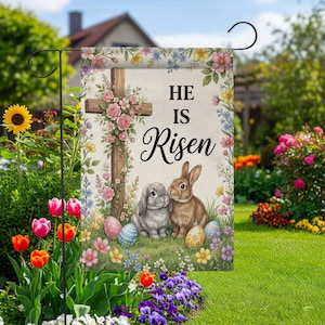 Könnte beinhalten: Eine Gartenflagge mit einem Holzkreuz, das mit rosa Blumen geschmückt ist und die Worte "HE IS Risen" trägt. Zwei Hasen sitzen in einem Feld mit Ostereiern. Die Flagge ist von bunten Blumen und Grün umgeben.