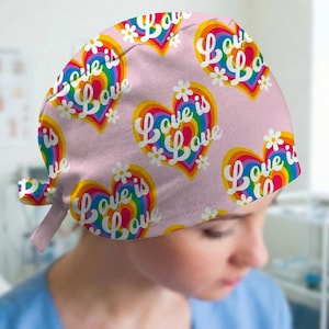 May include: A pink scrub cap featuring a repeating pattern of rainbow-colored hearts and white daisies. The text "Love is Love" is printed in white script across each heart. The cap has a tie-back closure and a rounded crown design.